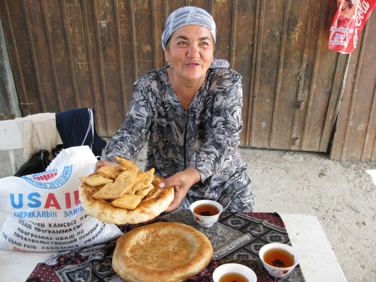 Женщина угощает боорсоками и лепешкой. Мешок муки с логотипом USAID лежит рядом.