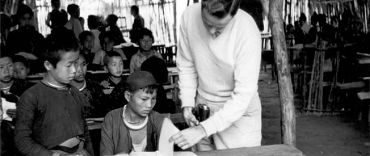 Meo tribe children and Hal Freeman, right, look at textbooks provided by USAID in Laos. In the background, a parachute serves as